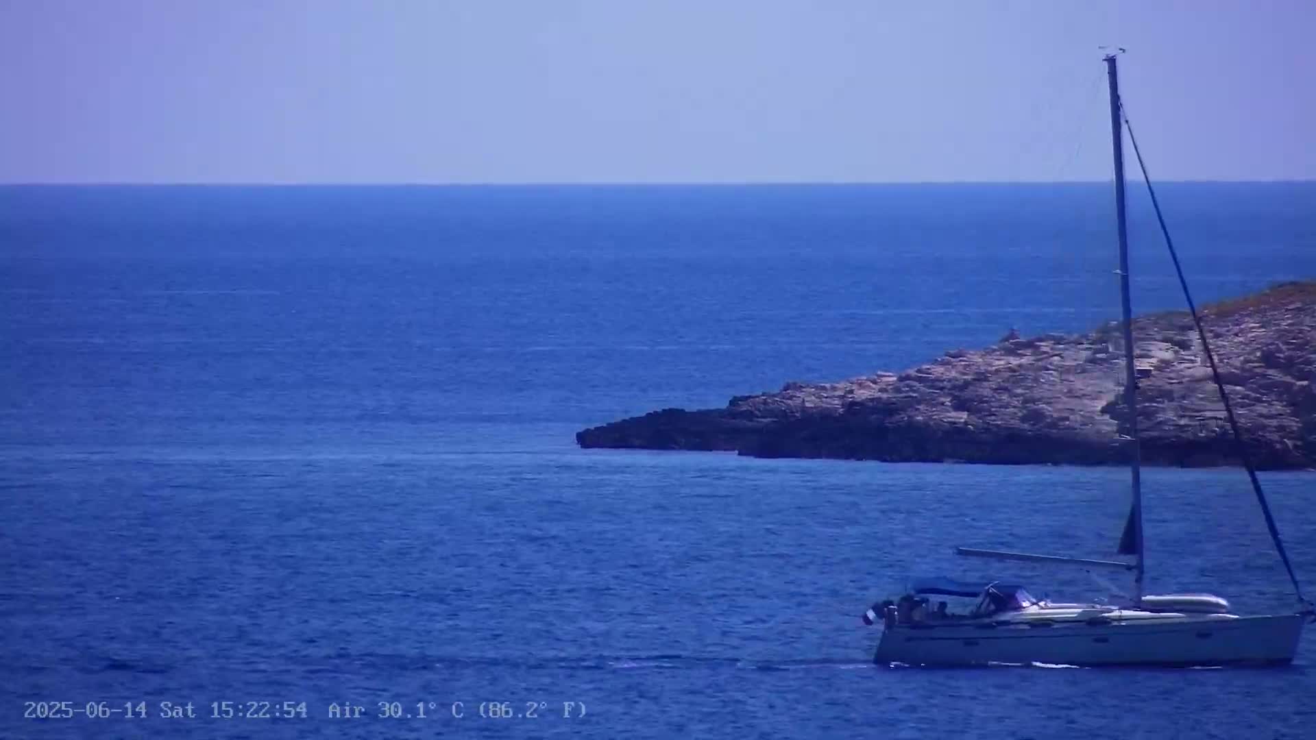 A sailboat is anchored in a calm, blue sea near a rocky outcrop on a clear, sunny day.