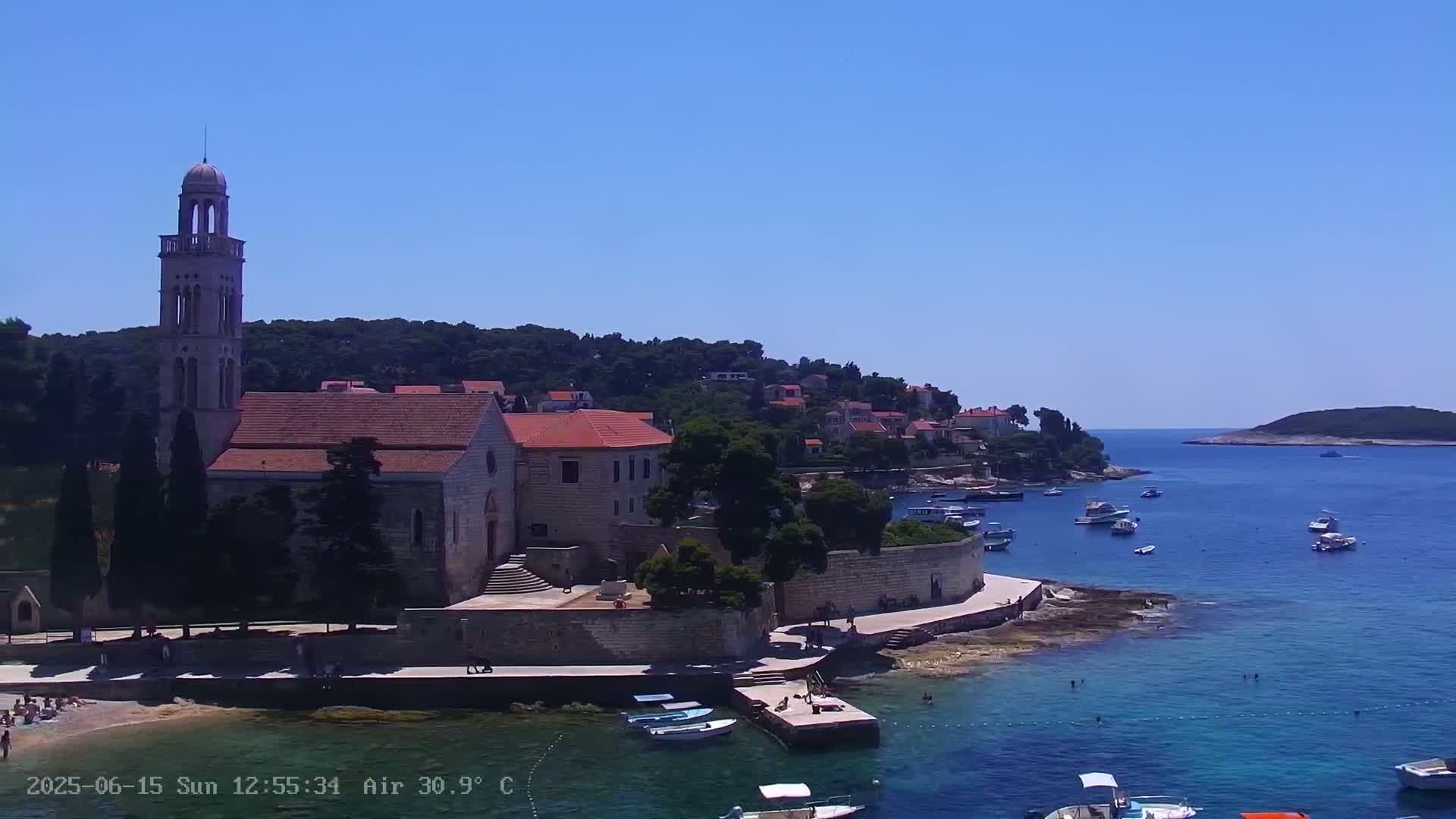 A stone church with a tall bell tower sits on a small peninsula overlooking a calm, blue sea with several boats and people swimming, under a clear blue sky.