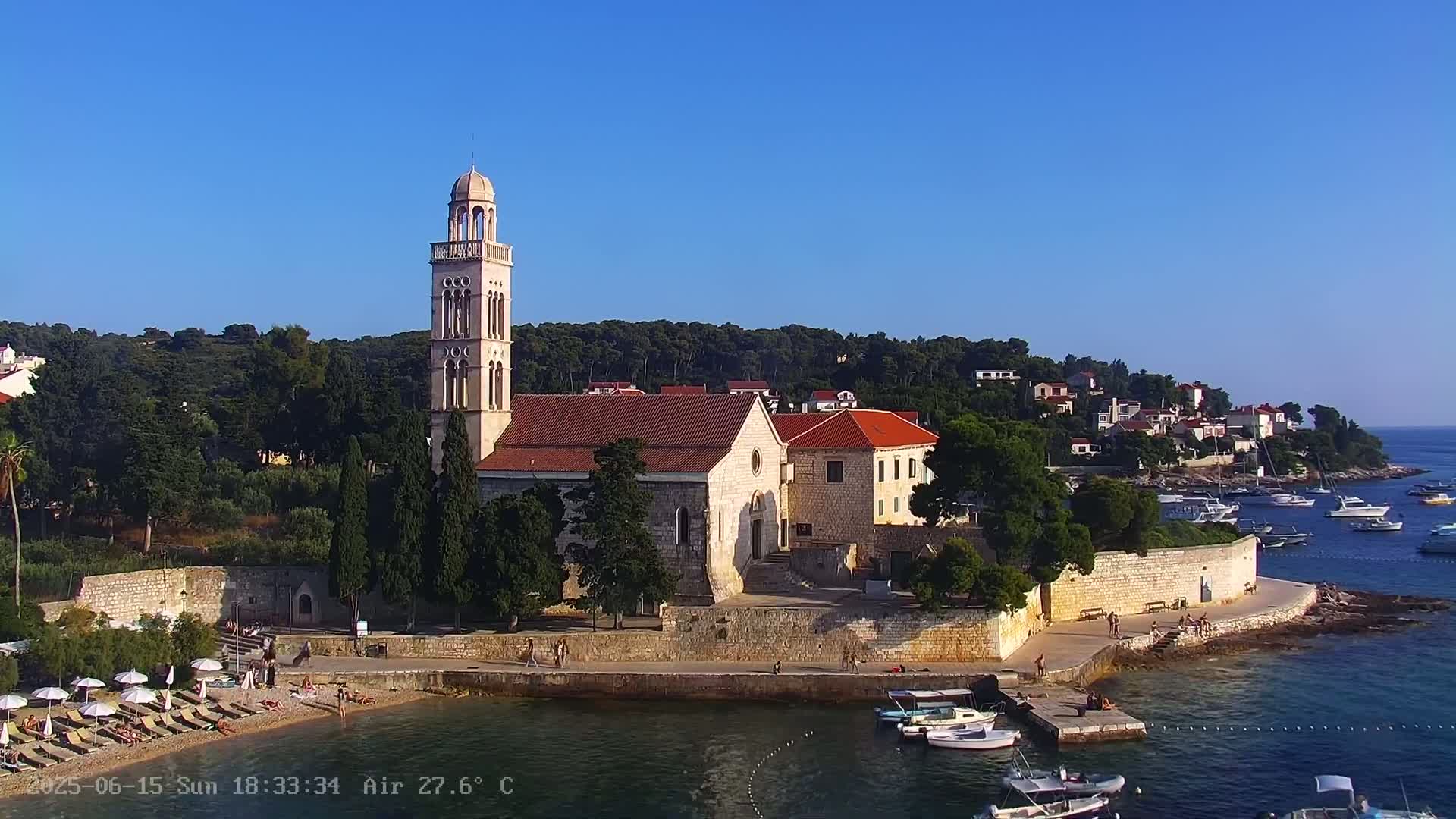 A church with a tall bell tower and red roof sits on a rocky coast, under a clear blue sky on a sunny day, with several boats in the calm water and people relaxing on a nearby beach.