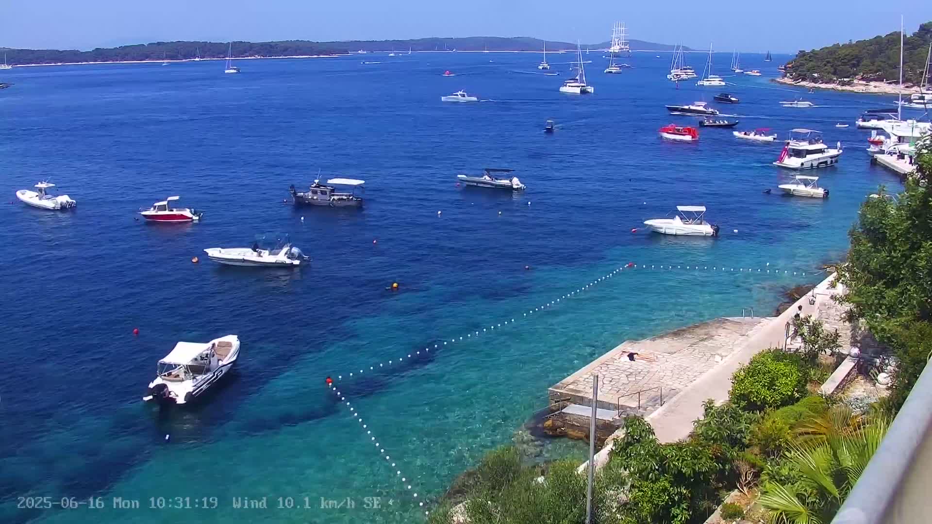 A sunny day shows a calm, clear blue sea with numerous boats of various sizes, some anchored, others underway, near a rocky shoreline with a paved area and lush vegetation.