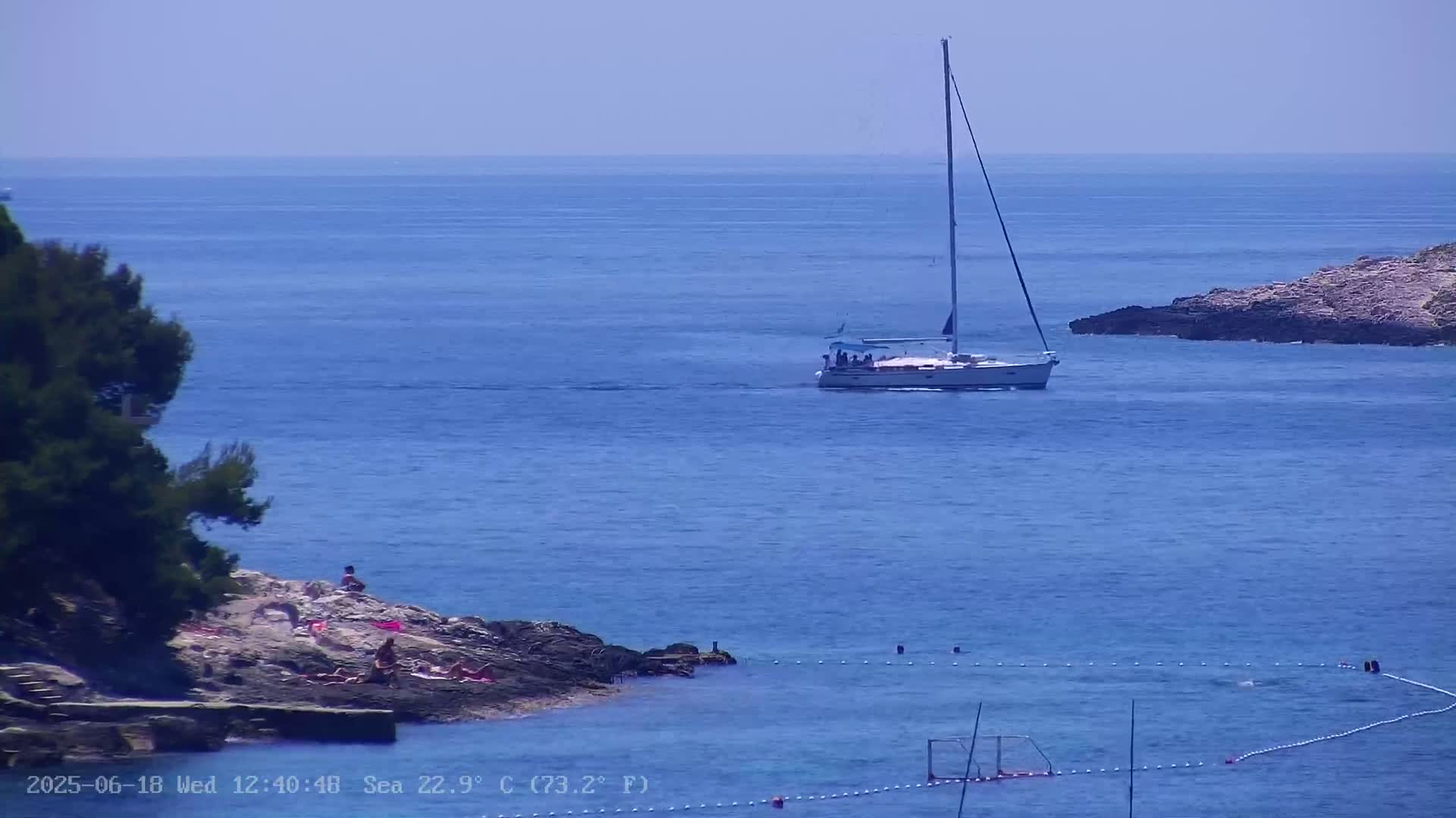 A sailboat is on a calm blue sea near a rocky shore with several people sunbathing, and a water polo goal is visible in the water.