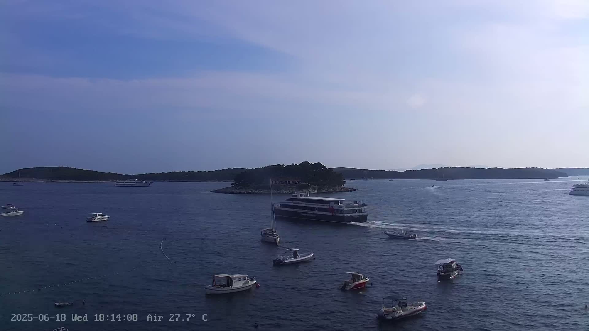 A calm, sunny day shows a body of water with several small boats and a large ferry near a small, tree-covered island and larger landmasses in the distance.