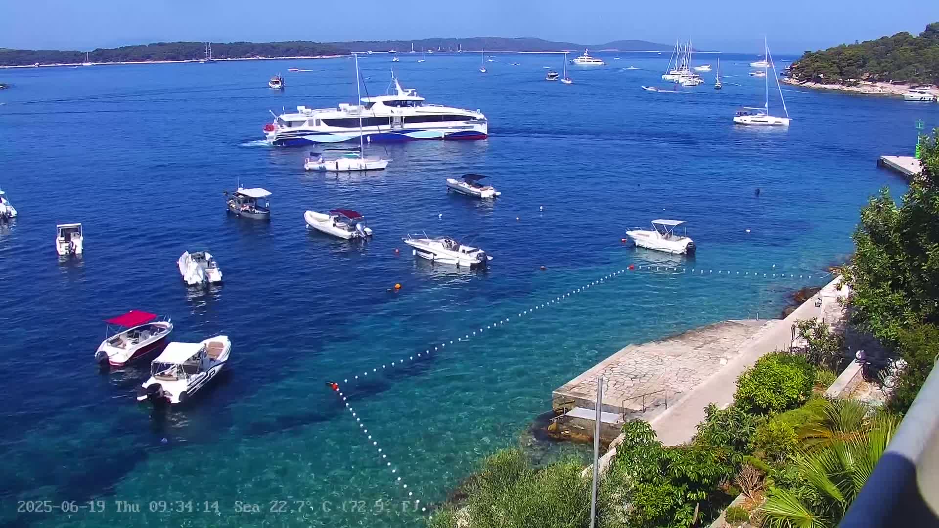 Numerous boats and yachts are anchored in a calm, clear blue sea on a sunny day, with a shoreline featuring vegetation and a paved area.