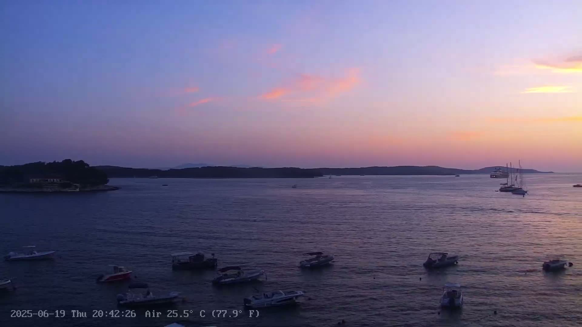 Numerous small boats are moored in calm water at sunset under a pastel-colored sky near a small island and distant shoreline.