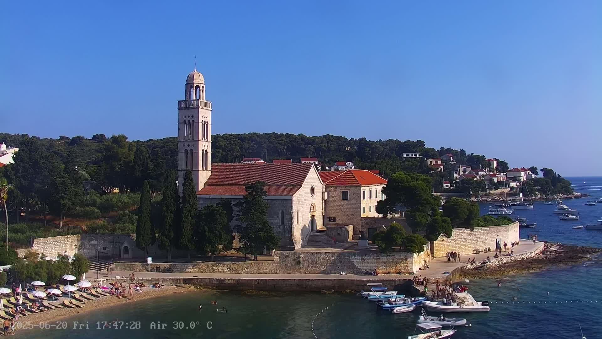 A church with a tall bell tower sits on a rocky coast overlooking a calm bay with several boats and people relaxing on a nearby beach on a sunny day.