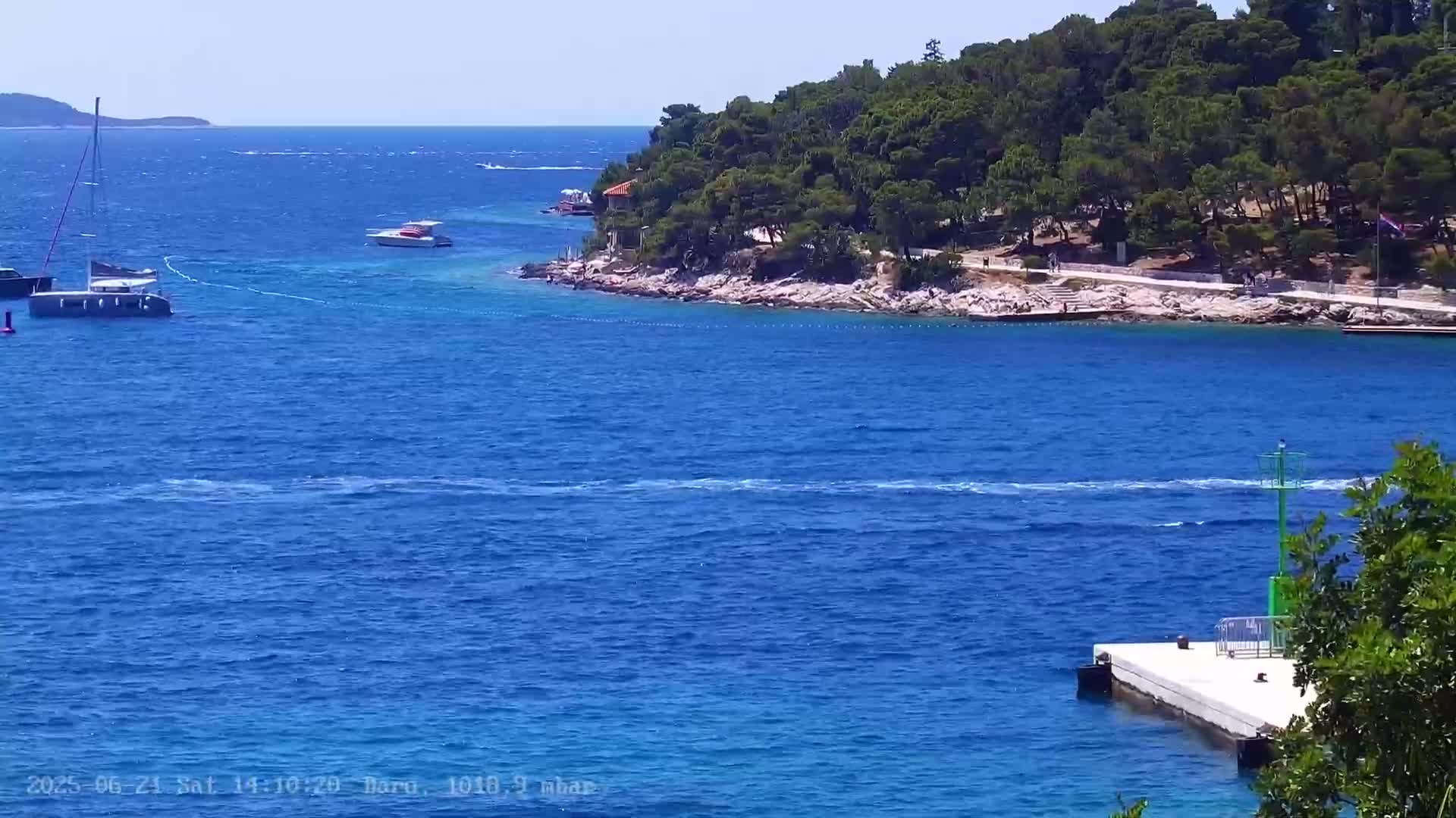 A sunny day shows a calm, blue sea with several boats near a small, rocky island covered in green trees.