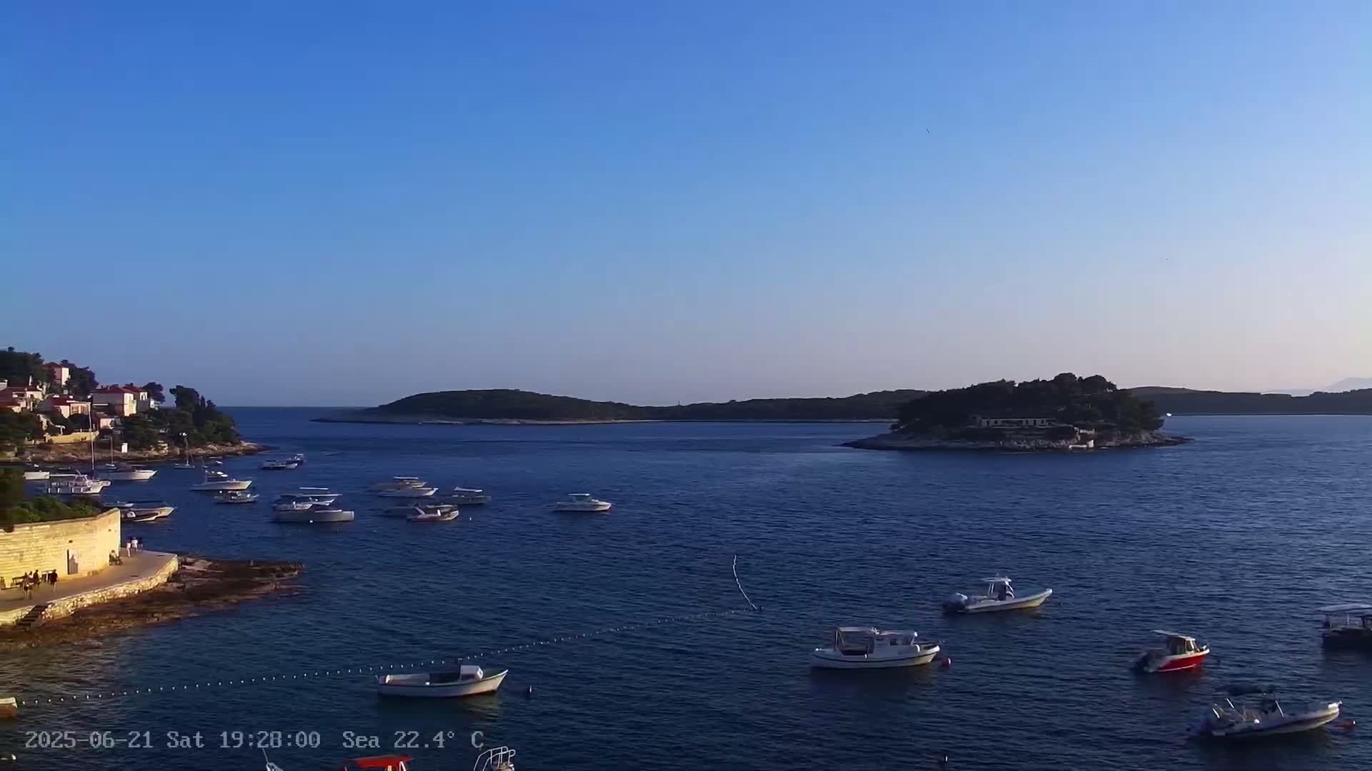 A calm, clear day reveals numerous boats moored in a bay near a small island and a shoreline with buildings.