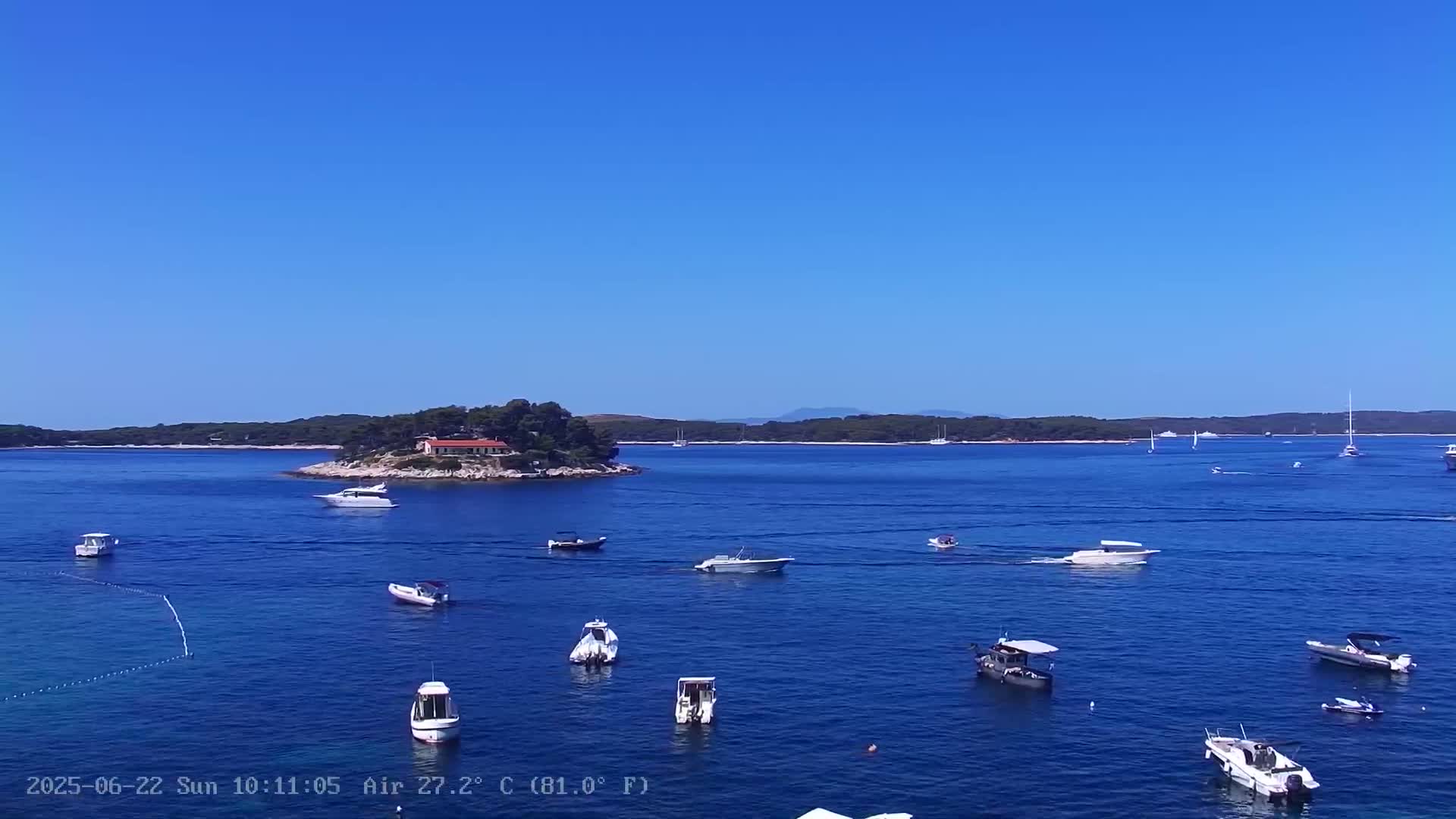A small island with a house sits in a calm, blue sea dotted with numerous boats under a clear, bright blue sky.