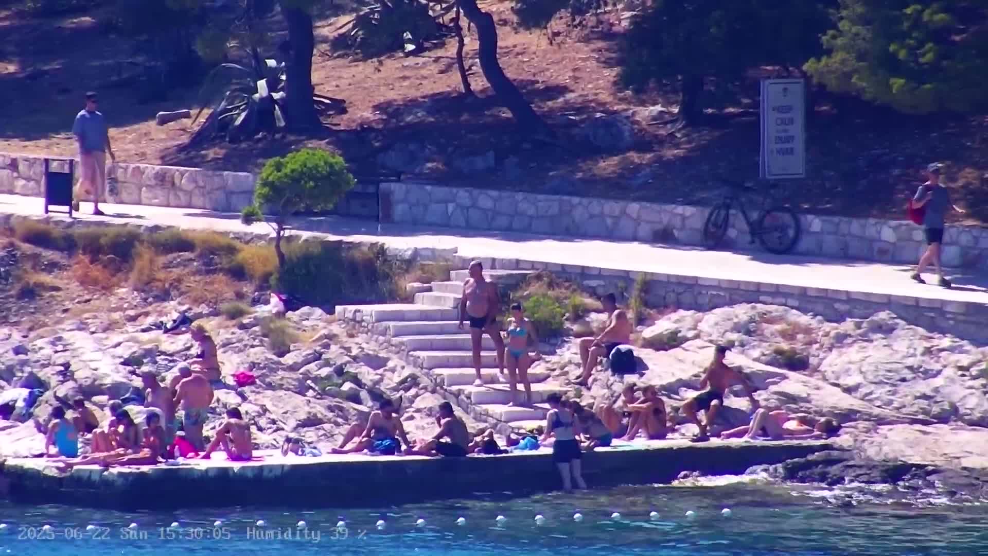 Many people are sunbathing on rocks and a low wall by the water on a sunny day.