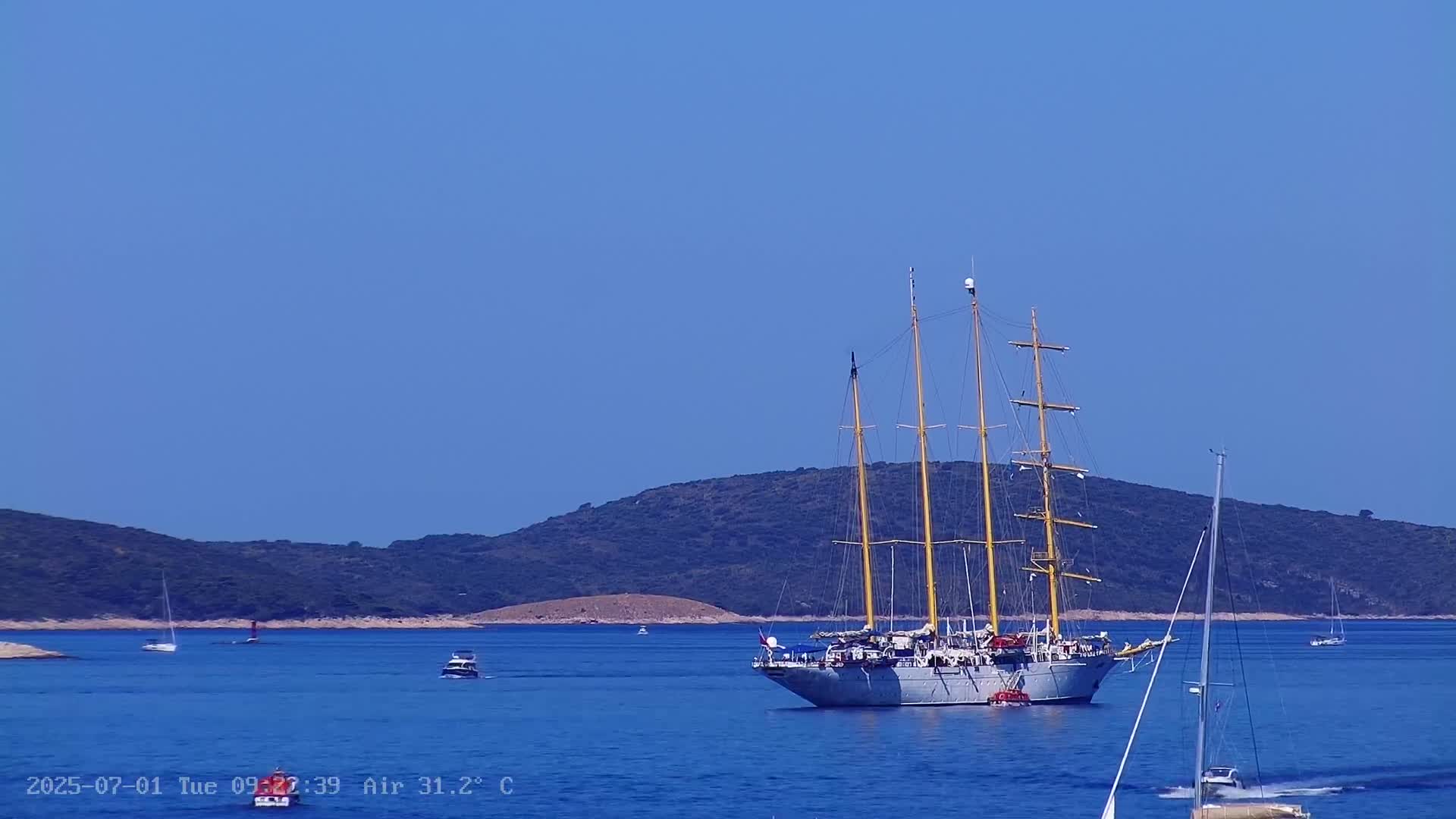 A large sailing ship is anchored in a calm, blue sea near a green, hilly island under a clear blue sky.