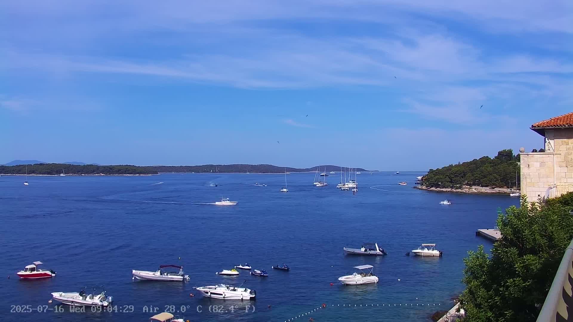 A sunny day shows numerous boats and yachts in a calm bay, with islands in the distance and a stone building partially visible in the foreground.