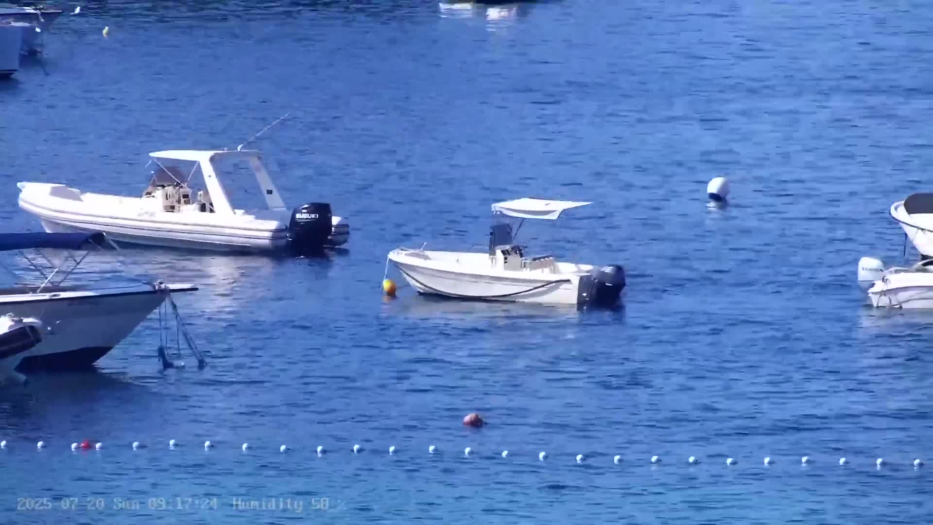 Several small motorboats are moored in calm, blue water, with a line of white buoys visible in the foreground.