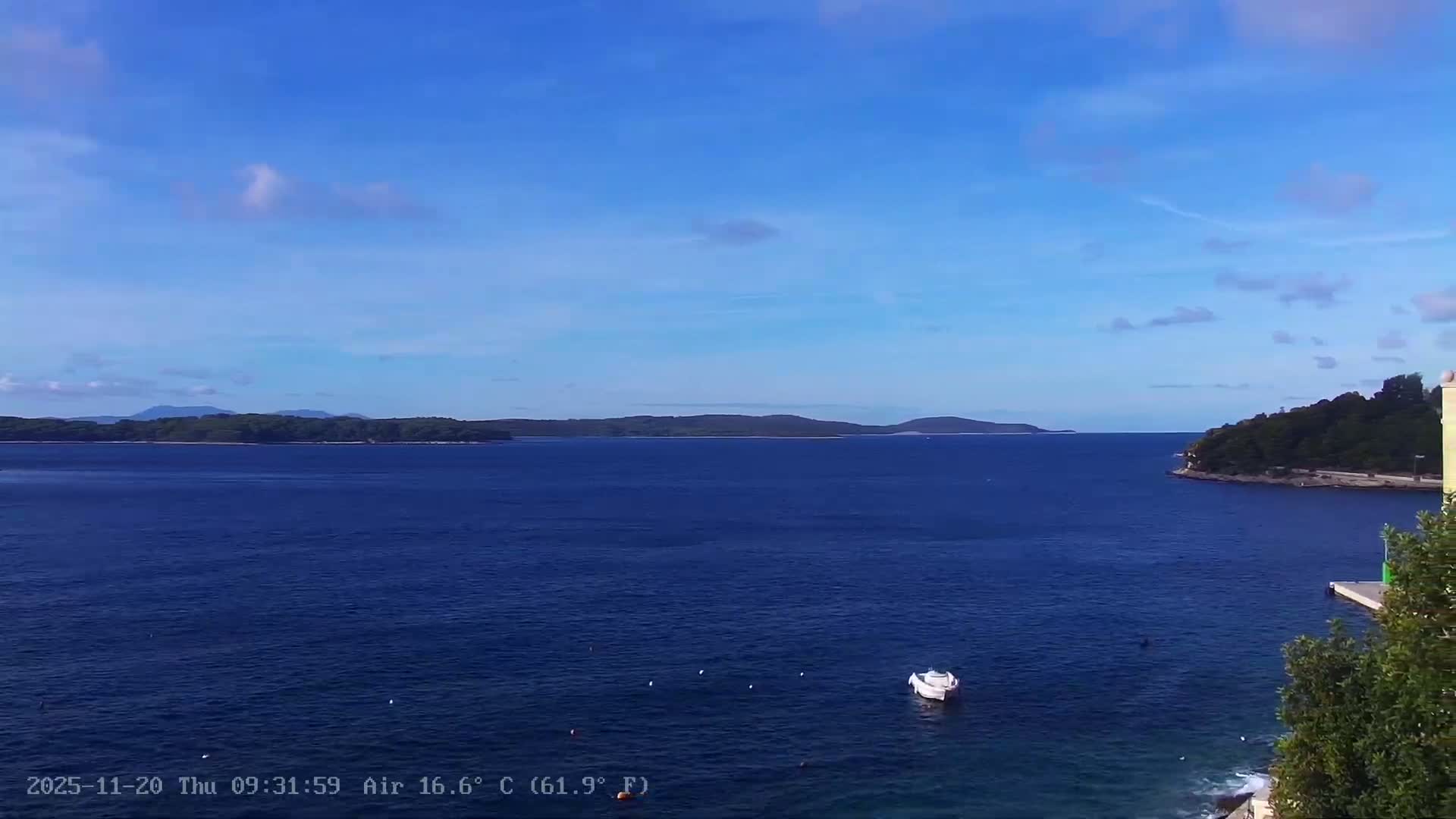A sunny, partly cloudy day illuminates a tranquil deep blue sea with a small white boat and buoys, bordered by distant forested islands and a green coastline with a building partially visible on the right.