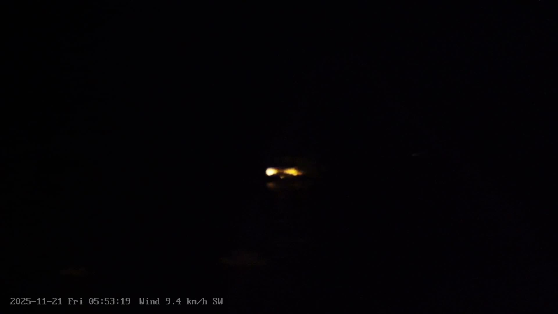 A sunny, partly cloudy day illuminates a tranquil deep blue sea with a small white boat and buoys, bordered by distant forested islands and a green coastline with a building partially visible on the right.