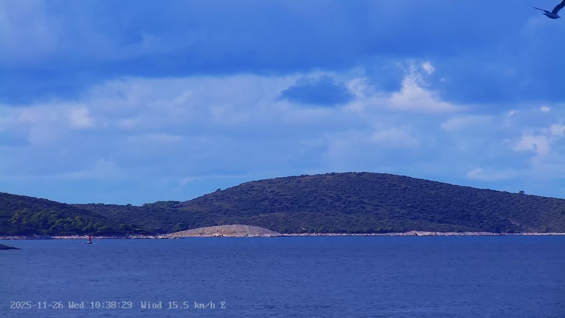 A calm, deep blue sea is framed by verdant, tree-covered islands under a partly cloudy sky, with a small red-sailed boat in the mid-ground and a bird flying overhead on a bright day.