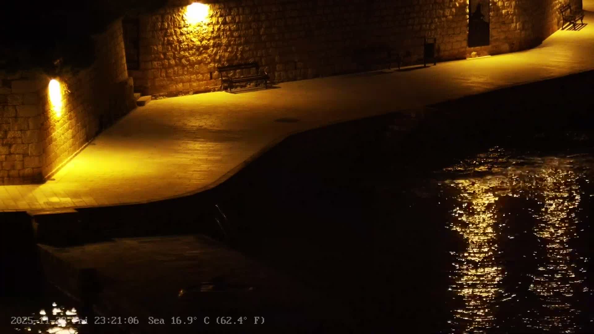 A warmly lit stone walkway with benches lines a dark body of water reflecting shimmering light at night under clear skies.