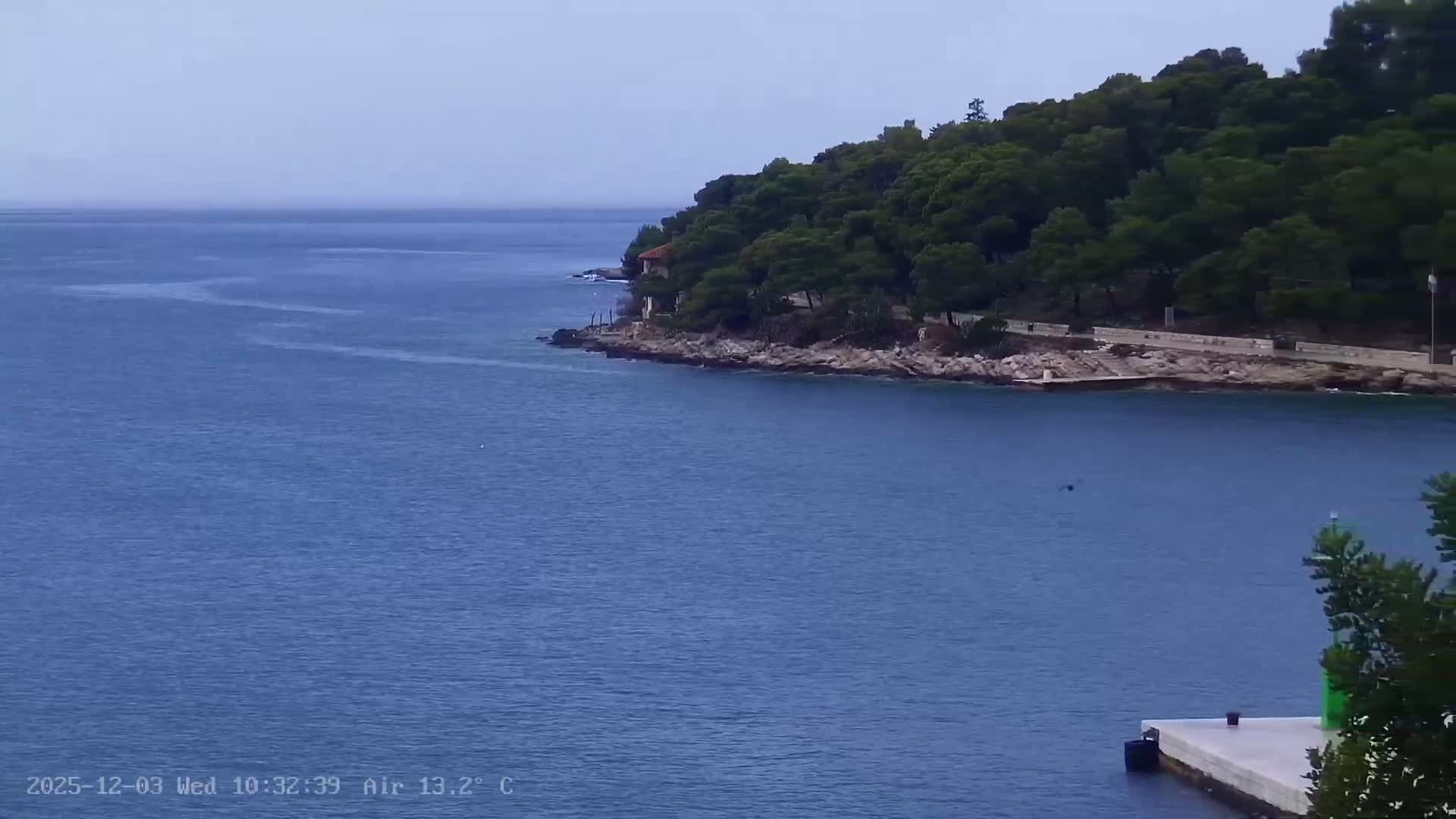 On an overcast day, a calm blue sea stretches towards the horizon, bordered on the right by a tree-covered coastline featuring a small house and a paved pathway, with a white concrete pier and green navigational marker visible in the bottom right.