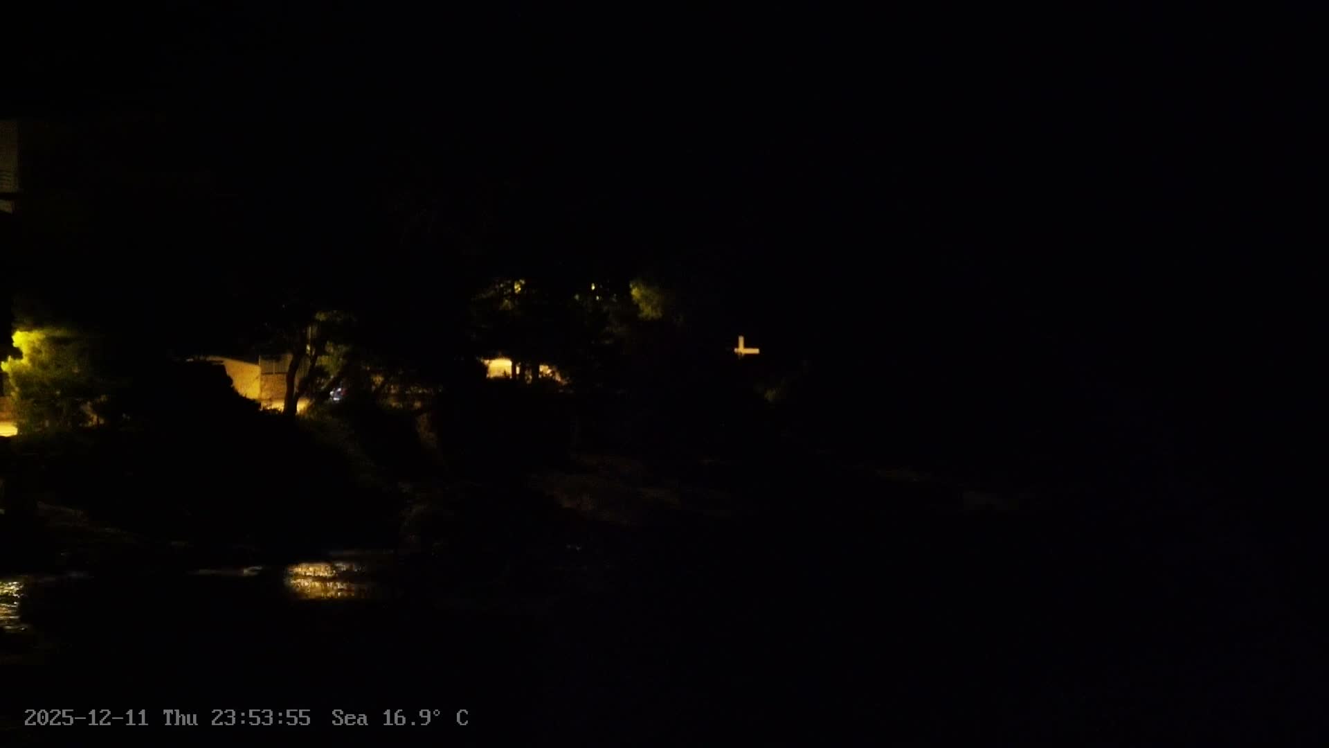 An extremely dark night scene reveals dimly lit buildings and trees along a sea shoreline, with light reflections on the water surface, under clear weather conditions.