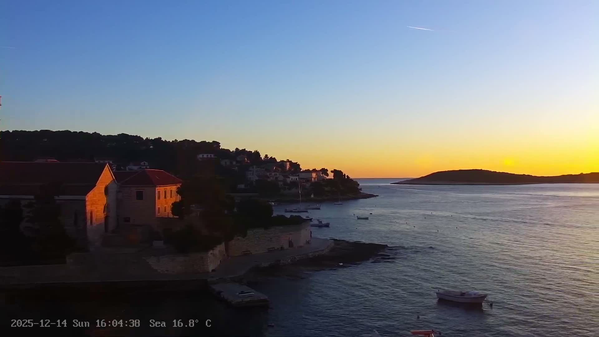 An extremely dark night scene reveals dimly lit buildings and trees along a sea shoreline, with light reflections on the water surface, under clear weather conditions.