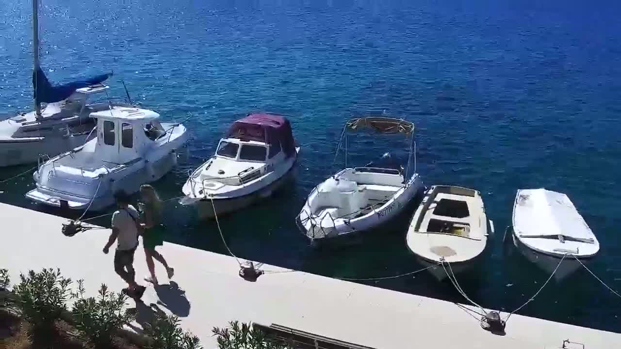 A couple walks past several small motorboats moored at a dock on a sunny day with calm, blue water.