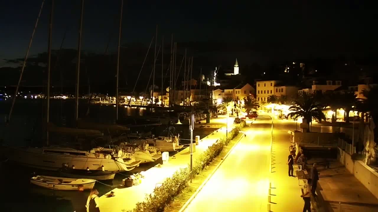 A brightly lit waterfront promenade at night, lined with docked sailboats and palm trees, leads to a village with buildings illuminated under a dark sky.