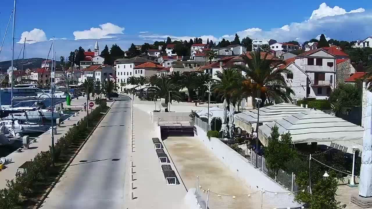 A sunny day shows a waterfront town with boats docked at a marina, a paved road lined with palm trees leading to a town square with buildings and an outdoor bocce ball court.