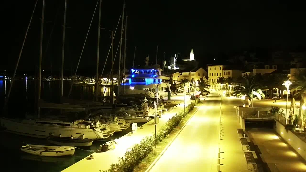A brightly lit marina at night, filled with sailboats and a larger vessel, overlooks a paved waterfront promenade lined with palm trees and buildings.