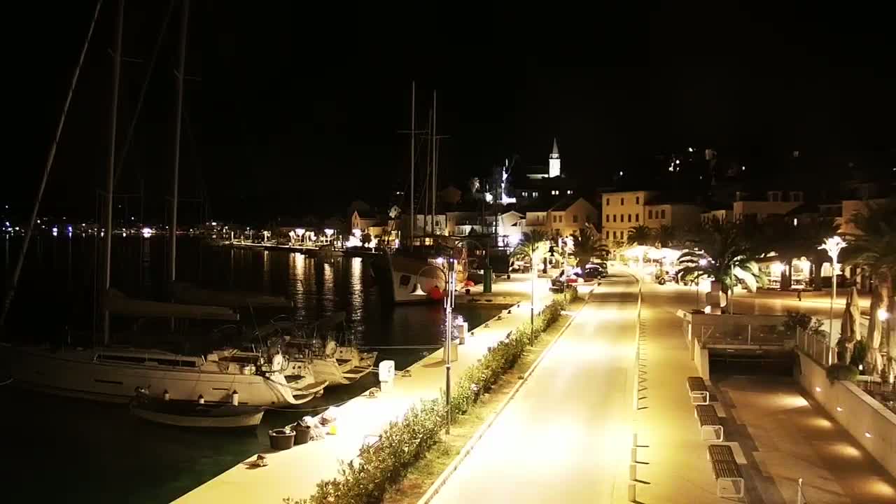 A nighttime view of a harbor town with sailboats docked, a waterfront promenade lined with lights and palm trees, and buildings illuminated along the shoreline.