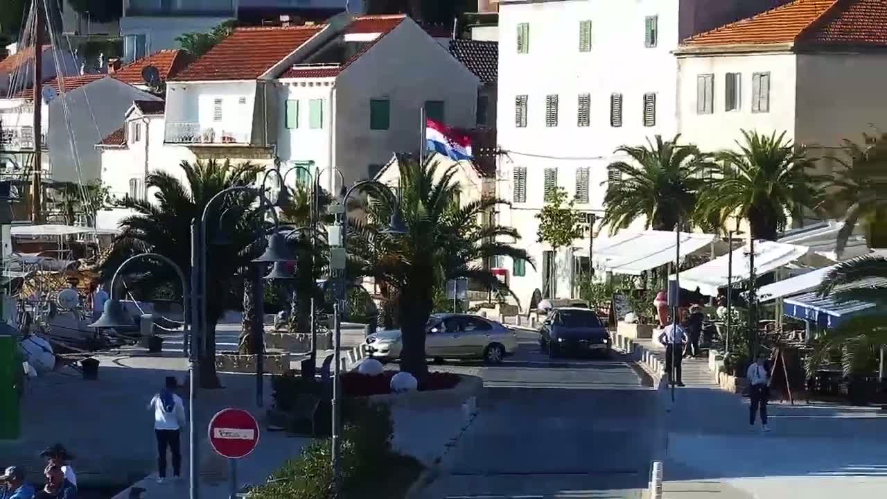 A sunny day reveals a Mediterranean-style waterfront town with white buildings, palm trees, outdoor cafes, and a few cars parked along the street.