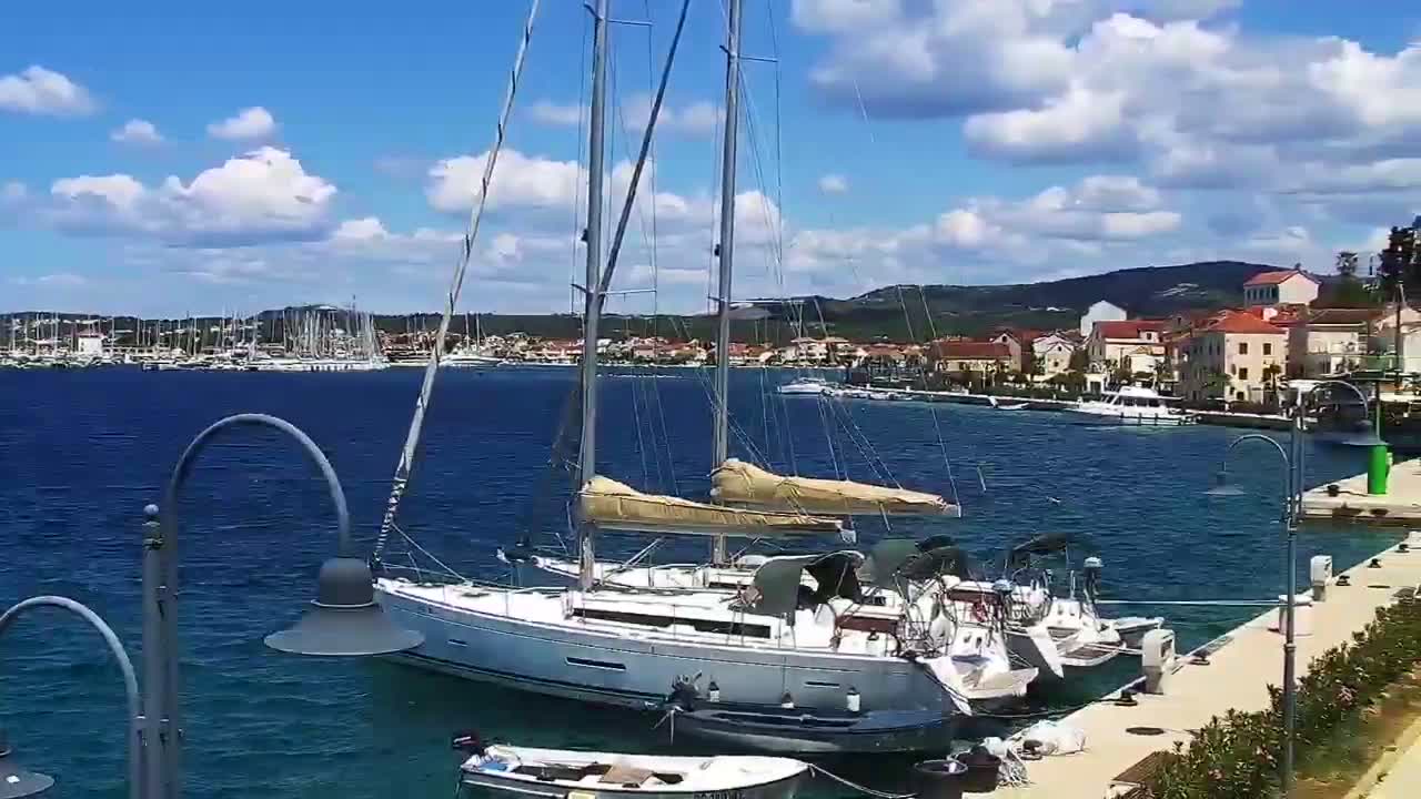 A large sailboat is docked at a marina on a sunny day with partly cloudy skies, numerous other boats are visible in the harbor and a town is in the background.