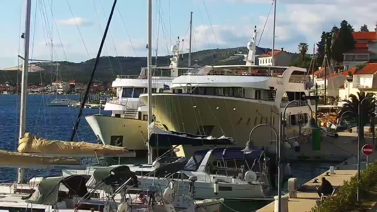 Several yachts and sailboats are docked at a marina on a sunny day, with a small town and hills visible in the background.
