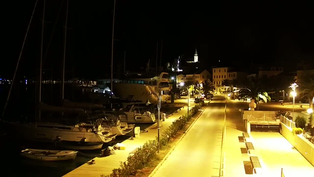 A nighttime view of a harbor with sailboats and a larger vessel docked, adjacent to a town with illuminated streets and buildings.