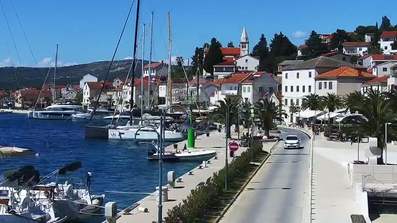 A waterfront town with numerous sailboats docked in a harbor, under sunny skies, features a road leading through the town.