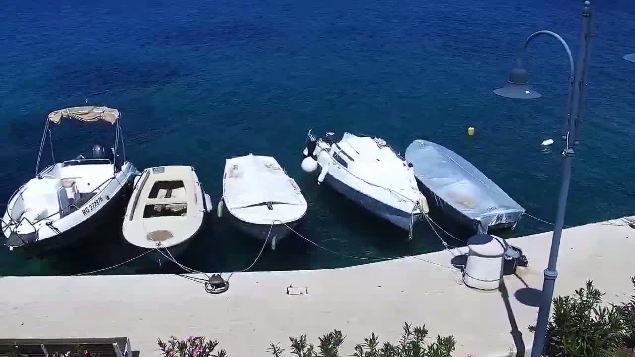 Four small boats are moored along a light-colored dock beside calm, clear blue water on a sunny day.