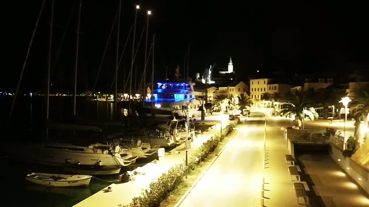 A nighttime view of a harbor filled with sailboats and a larger yacht, with a brightly lit waterfront promenade leading to a village under a clear, dark sky.