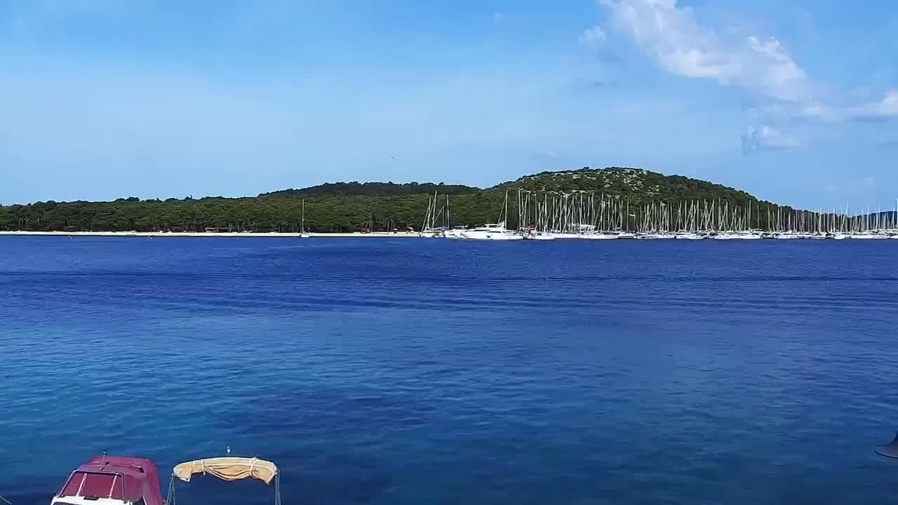 A calm, blue sea stretches towards a tranquil shore lined with numerous sailboats and a verdant, hilly island under a partly cloudy sky.