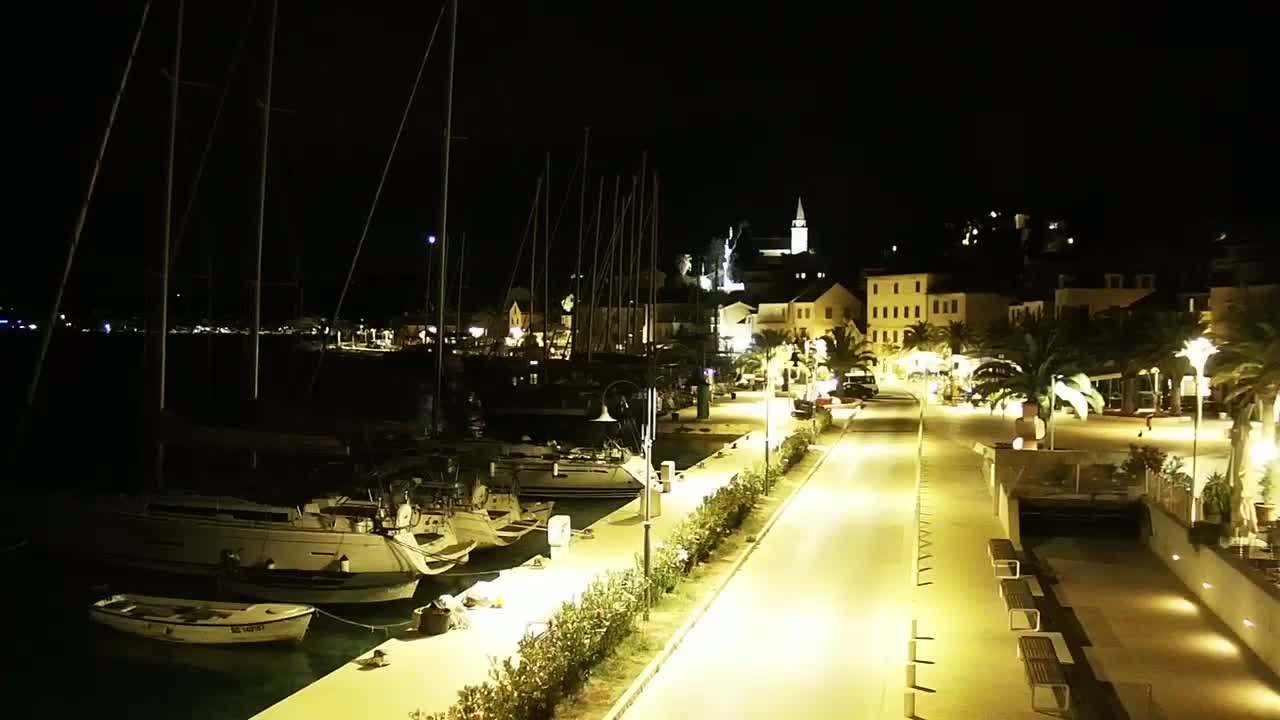 A nighttime view of a harbor filled with sailboats, a waterfront promenade with palm trees and streetlights, and a small town with buildings lit up in the background.