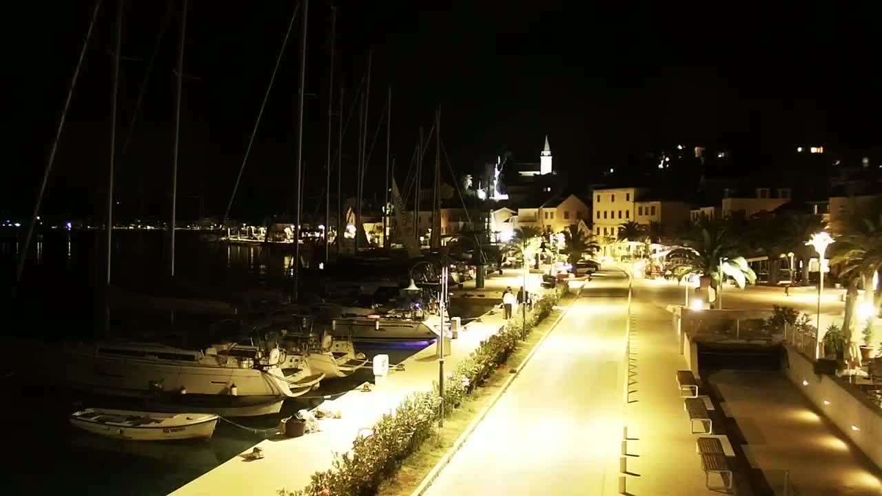 A nighttime view of a harbor filled with sailboats, a waterfront promenade lined with palm trees and benches, and a small town illuminated by streetlights.