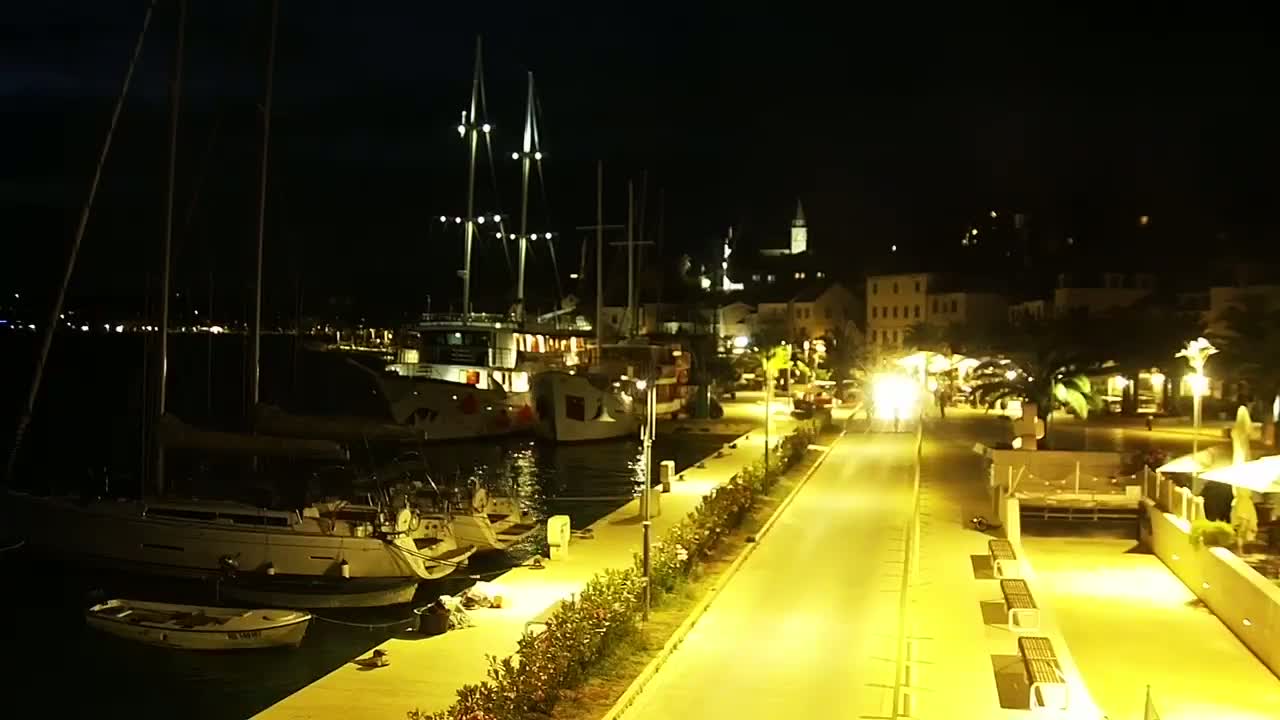 A nighttime view of a harbor with sailboats and a waterfront walkway illuminated by streetlights, overlooking a town.