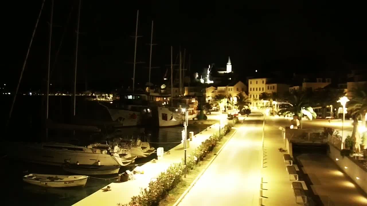 A nighttime view of a harbor with sailboats docked alongside a brightly lit waterfront promenade leading to a small town.