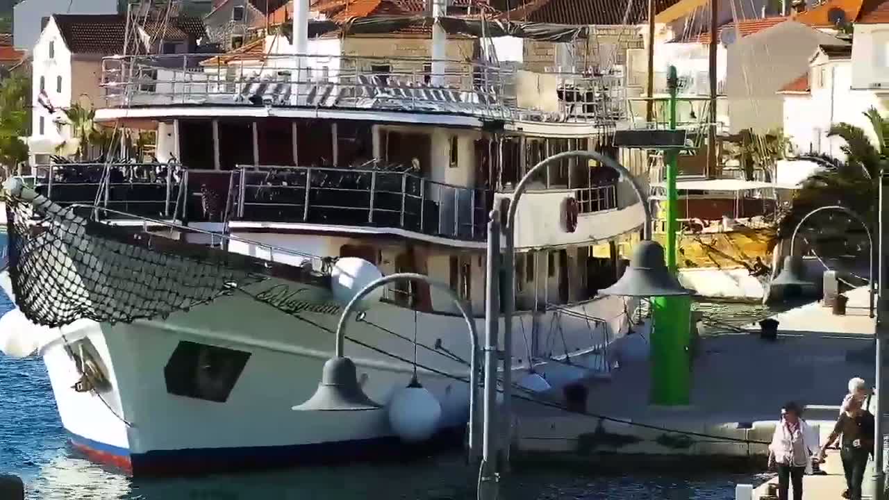 A large passenger ship is docked at a waterfront, and a couple strolls along the nearby quay on a sunny day.