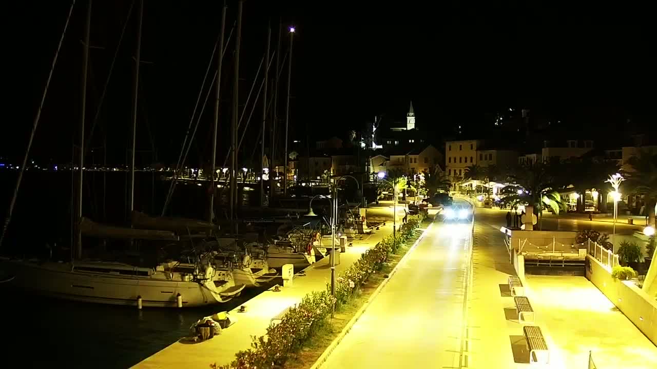 A nighttime harbor scene shows sailboats docked along a waterfront promenade, illuminated by streetlights, with a town visible in the background under a clear sky.