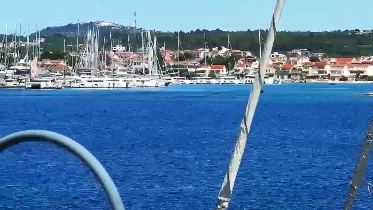 A marina filled with numerous sailboats is seen from a boat on a sunny day with calm, blue water.