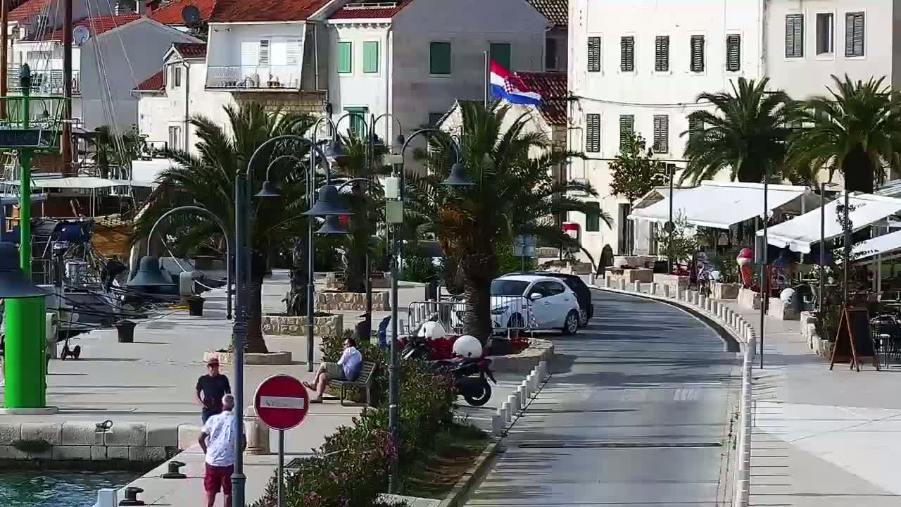 A sunny seaside town square with palm trees, parked cars, people relaxing, and boats moored in the harbor.