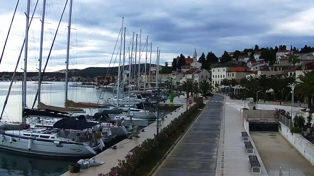 A waterfront promenade with numerous sailboats docked alongside, under a cloudy sky, overlooks a town with buildings and palm trees.