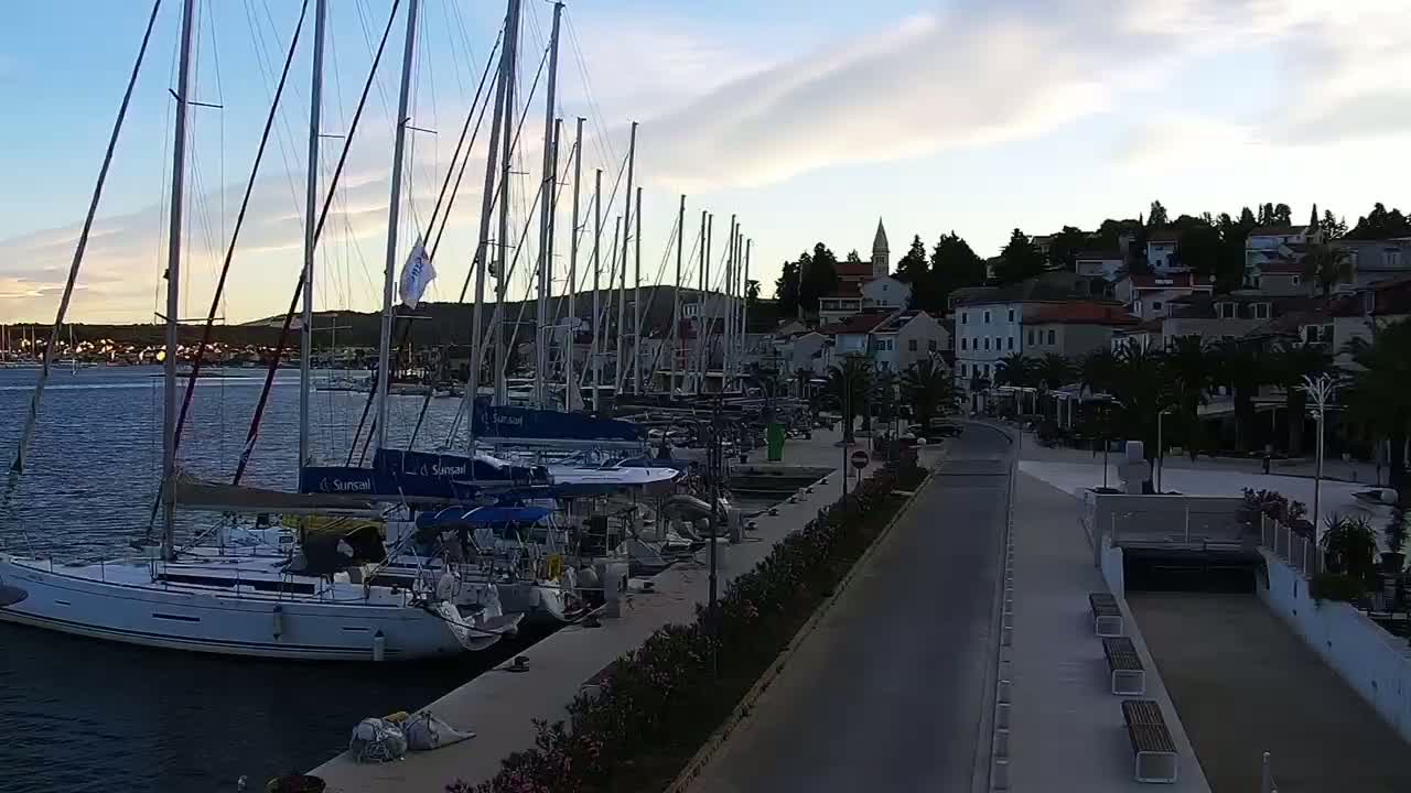 Many sailboats are docked at a marina next to a waterfront town under a partly cloudy twilight sky.