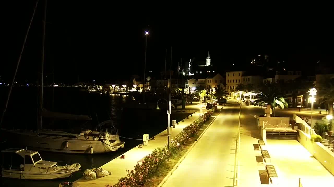 A nighttime view of a harbor town's waterfront, showing boats moored at a dock and a paved walkway lined with lights leading to buildings illuminated against a dark sky.