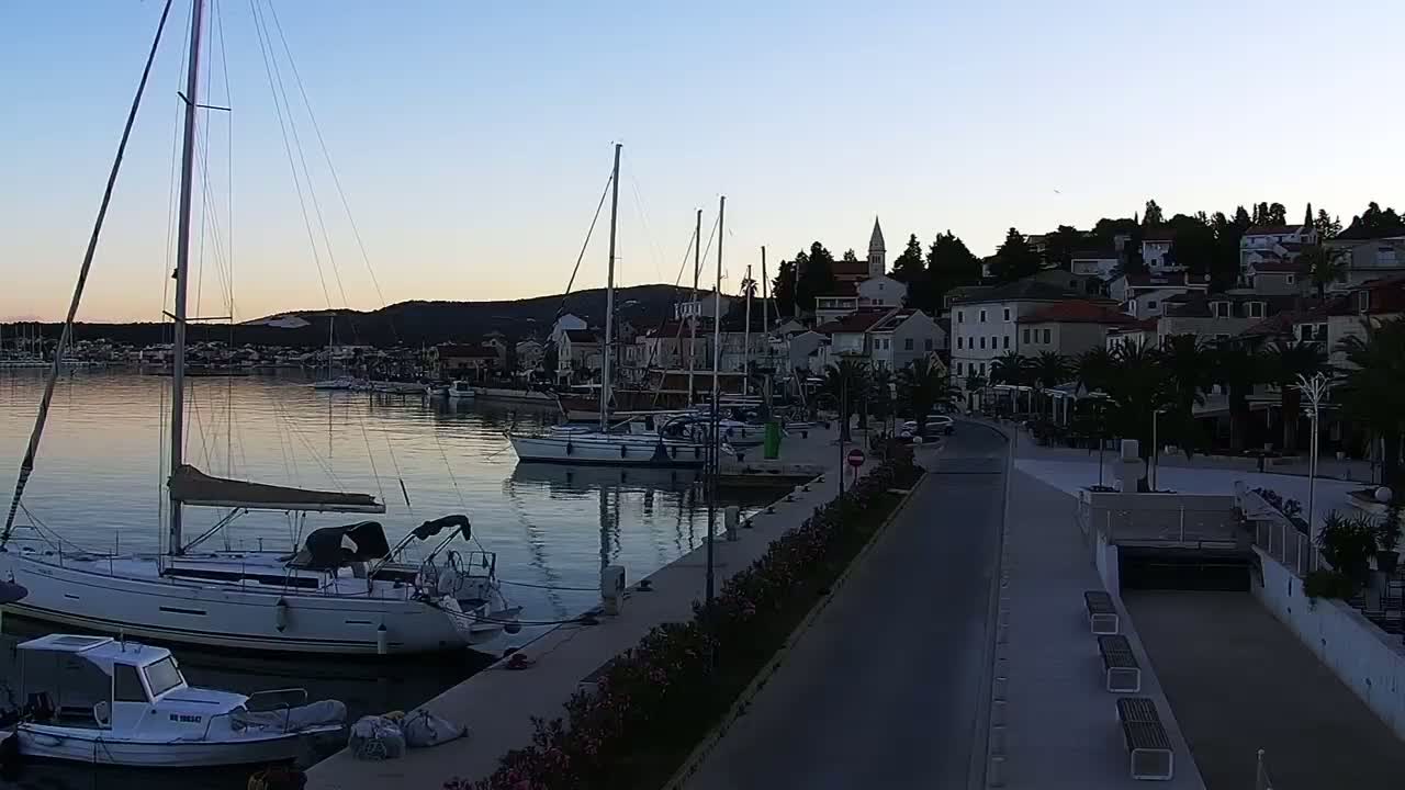 A calm harbor at dawn, filled with sailboats moored alongside a waterfront promenade lined with buildings and palm trees.
