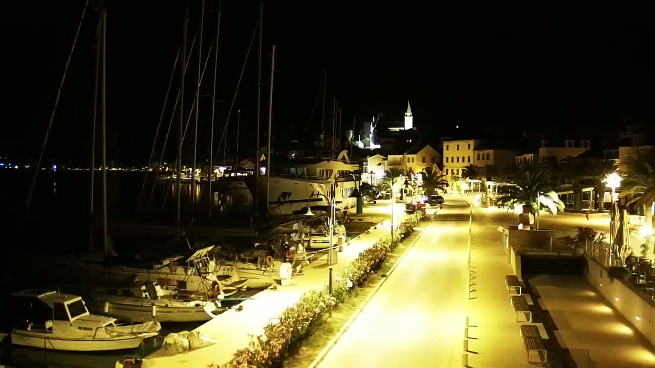 A nighttime view of a harbor with docked sailboats and yachts, leading to a brightly lit waterfront town under a clear, dark sky.