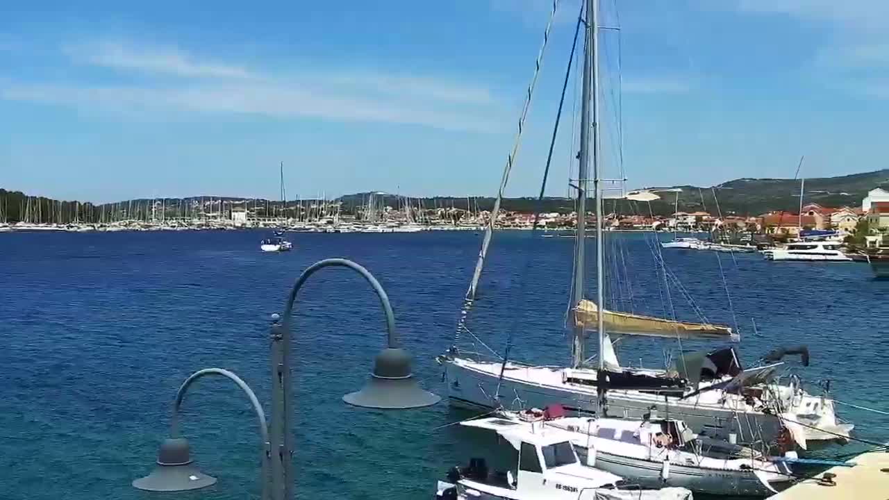 Two sailboats are moored in a calm, blue harbor on a sunny day, with a town and numerous other boats visible in the background.